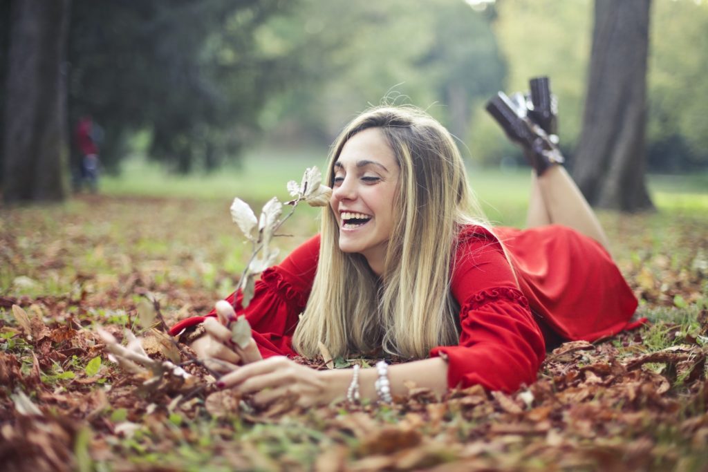 Selective Focus Photo of Laughing Woman in Red Dress Lying on Her Stomach on Dry Leaves While Holding a Dry Branch